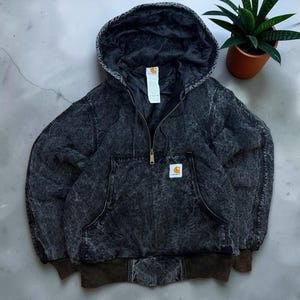 Könnte beinhalten: Eine schwarze, säuregewaschene Kapuzenjacke mit Kängurutasche, halbem Reißverschluss und braunem Rippsaum. Die Jacke hat ein Carhartt-Markenlogo auf der Tasche und ein schwarzes Steppfutter mit einem weißen Markenetikett am inneren Kragenbereich.
