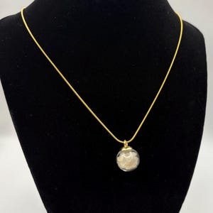 May include: A gold-colored necklace with a delicate chain and a clear glass pendant. The pendant is a small sphere filled with a cluster of white, fluffy material. The necklace is displayed on a black velvet bust.