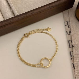 May include: A gold-colored bracelet with a double-strand beaded chain and a single chain link. The bracelet features a circular design in the center and a heart-shaped charm near the clasp. The jewelry is displayed on a white surface.
