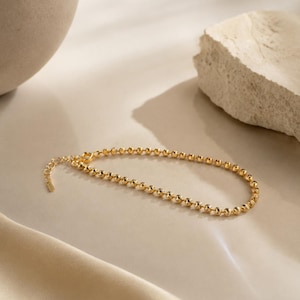 May include: A gold-toned chain bracelet composed of small, textured spherical beads linked together. The bracelet features a short extender chain with a small rectangular tag at the end, showcasing a polished and reflective metallic finish.