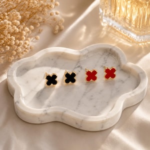May include: Four pairs of clover-shaped stud earrings in black and red, displayed on a white marble tray. The earrings have gold-colored posts and are arranged in a row. The tray is cloud-shaped and sits on a satin surface.
