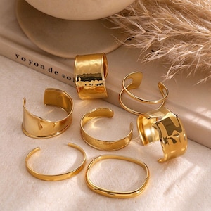 May include: A collection of seven gold-toned metal cuff bracelets and rings. The pieces feature various widths and textures, including smooth polished surfaces and hammered finishes. Each item is an open-band design, showcasing a reflective metallic sheen.