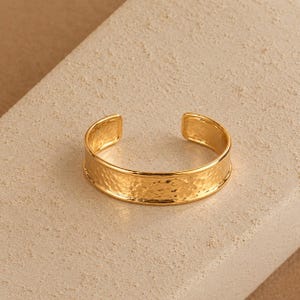 May include: A gold-toned open cuff bracelet featuring a hammered, textured surface on the exterior band. The piece has a polished, smooth interior and rounded edges, forming a circular shape with a gap at the back for adjustment.