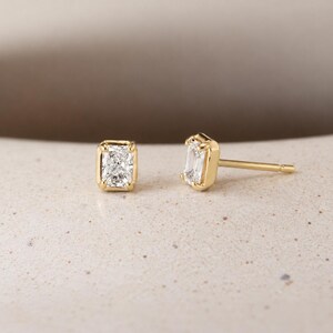 May include: A pair of stud earrings featuring rectangular radiant-cut clear stones set in polished yellow gold. One earring is shown from the front, while the other is positioned to display the side profile and the gold post backing.
