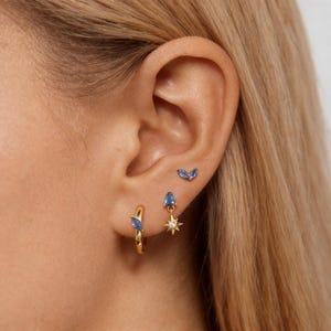 May include: Three gold-tone earrings featuring blue marquise and pear-cut stones. One small hoop earring with a blue stone, one stud earring with a pear-cut blue stone and a starburst charm, and one small stud earring with two blue marquise stones.
