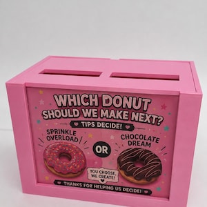 May include: A pink rectangular tip jar with two slots on top. The front features a sign reading: WHICH DONUT SHOULD WE MAKE NEXT? TIPS DECIDE! SPRINKLE OVERLOAD OR CHOCOLATE DREAM. YOU CHOOSE, WE CREATE! THANKS FOR HELPING US DECIDE! with donut graphics.