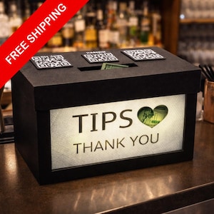 May include: A black tip box with a rectangular shape. The front of the box has the words "TIPS THANK YOU" in white. A heart-shaped cutout reveals a bill. Three QR code stickers are on top.