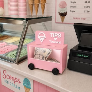 May include: A pink rectangular tip box shaped like a small cart with black wheels. The top sign features a white ice cream cone icon, the text "TIPS", a heart symbol, and the word "Thanks!". The box contains paper currency visible through a clear front panel.