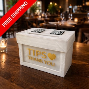 May include: A white tip box with the words "TIPS THANK YOU" in gold lettering and a gold heart. The box has two QR codes on top and is on a wooden table. The background is a restaurant setting.