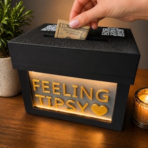 May include: A black rectangular tip box with a slot on top for currency. The front features illuminated gold lettering that reads FEELING TIPSY followed by a heart symbol. Two QR codes are printed on the top surface near the insertion slot.
