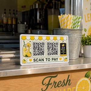 May include: A rectangular white sign with a yellow striped awning graphic, two QR codes, and a small chalkboard illustration reading "When life gives you lemons, make lemonade." The sign features a lemon drink graphic and the text "SCAN TO PAY."