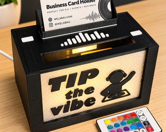 May include: A black rectangular tip box with a light-up front panel featuring the text "TIP the vibe" and a DJ graphic. It includes a top slot for business cards, a remote control for lighting, and a white power cord.
