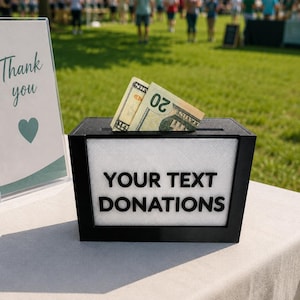 May include: A black rectangular donation box with a white front panel featuring the black text YOUR TEXT DONATIONS. A twenty-dollar bill is inserted into the top slot. Next to the box is a sign with the text Thank you and a heart icon.