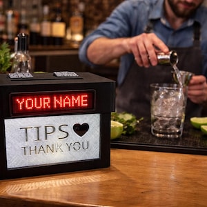 May include: A black tip jar with a red LED display that reads "YOUR NAME". The front of the jar says "TIPS ❤️ THANK YOU" in white lettering. A bartender is pouring a drink in the background.