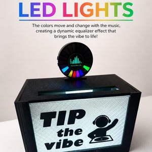 May include: A black rectangular tip box with a light-up front panel featuring the text "TIP the vibe" and a graphic of a DJ. A circular sound-reactive LED light display sits on top, showing a digital equalizer and the text "Music the Life".