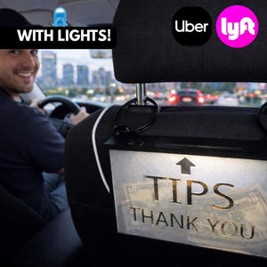 May include: A clear plastic tip box with the words "TIPS THANK YOU" in gold lettering, illuminated by a light. The box is attached to the back of a car seat. The image also includes the Uber and Lyft logos.
