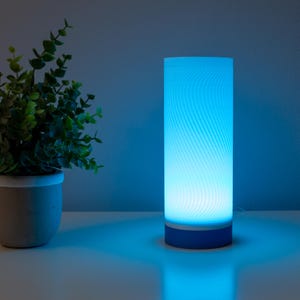 May include: A cylindrical blue LED lamp with a swirling pattern, emitting a soft glow. The lamp has a blue base and sits on a white surface. A potted plant with green leaves is to the left of the lamp.