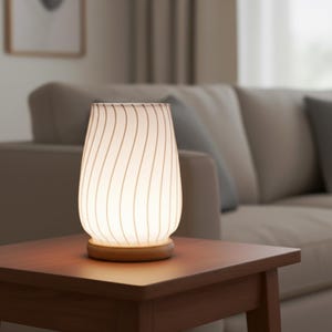 May include: A table lamp with a tapered, translucent white glass shade featuring vertical wavy lines. The lamp is supported by a circular, light-toned wooden base. The glass shade emits a soft, warm glow, illuminating the surrounding area.