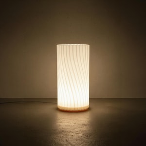 May include: A cylindrical table lamp with a translucent white shade featuring a vertical wavy ribbed texture. The lamp emits a warm, soft glow from within, illuminating the surface it rests upon while casting a gentle light in the surrounding area.