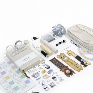 Peut inclure: Divers articles de papeterie, dont un carnet, des stylos, des autocollants et une trousse, sont disposés sur une surface blanche. Le carnet a une couverture beige et le texte "A Notebook". Sont également présents des éléments décoratifs comme une pince à cheveux en forme de fleur et un bijou de charme perlé.