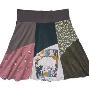 May include: A knee-length skirt featuring a wide dark gray waistband and a patchwork design. The panels include solid dark gray, dusty rose with a white floral print, dark teal, white with a graphic bear and leaf print, and olive green with a textured pattern.