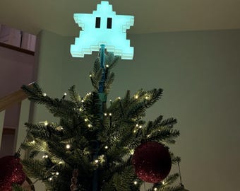 May include: A pixelated, light-blue star tree topper with two black eyes sits atop a decorated Christmas tree. The tree is adorned with small white lights and red and gold ornaments, creating a festive holiday scene.