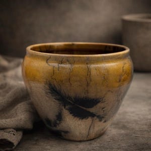May include: A handcrafted ceramic bowl with a wide rim and a tapered body. The bowl features a feather design in black against a beige and yellow background. The bowl is approximately 6 inches in diameter and 4 inches tall.