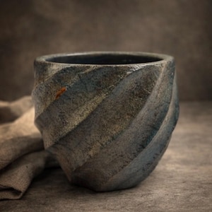 Könnte beinhalten: Eine handgefertigte Keramiktasse mit einem einzigartigen Spiralmuster. Die Tasse hat eine dunkle, gesprenkelte blau-graue Farbe mit subtilen Variationen. Der Innenraum ist dunkel, und die Tasse steht auf einer neutralen Oberfläche, was eine rustikale Ästhetik suggeriert.