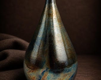 Puede incluir: Un jarrón de cerámica hecho a mano con forma de lágrima. El jarrón presenta un esmalte oscuro e iridiscente con tonos azules, bronce y dorado. El jarrón tiene un cuello estrecho y una base ancha y redondeada, de unos 25 cm de altura.