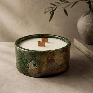 May include: A round candle in a textured, mottled green and brown ceramic vessel. The candle features white wax and two rectangular wooden wicks positioned in the centre. The exterior of the container has a weathered, earthy appearance with variations in colour tone throughout the surface.
