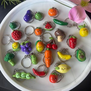 May include: A selection of handmade keyrings in the shapes of fruits and vegetables, such as a purple aubergine, green grapes, and a red strawberry. Each keyring has a silver ring. The keyrings are displayed on a white, circular tray.