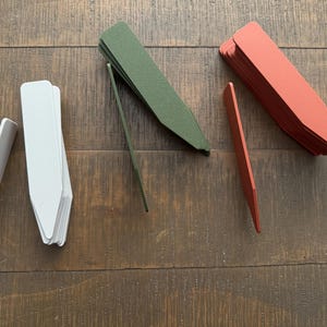 May include: A collection of plant markers in various colors, including white, green, and red-orange. The markers are made of a flat, rectangular material with a pointed end for insertion into the soil. They are arranged on a wooden surface.