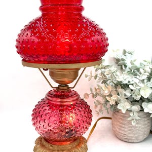 May include: A vintage red hobnail glass lamp with a ruffled top and a round base. The lamp has a gold-colored metal base and accents. The lamp is lit, showcasing its intricate hobnail texture. A decorative item for home decor.