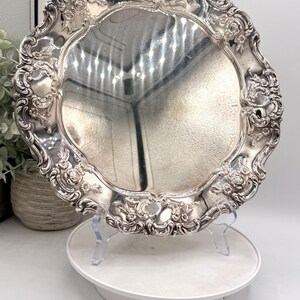 May include: A round silver-toned serving tray featuring an ornate, embossed border with intricate floral and scrollwork detailing. The central surface is smooth and reflective, while the edges are scalloped with a raised, decorative metal rim.