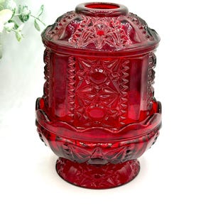 May include: A ruby red, vintage-style glass jar with a lid. The jar features intricate geometric patterns and a pedestal base. The lid has a decorative finial. The jar is likely used for storage or display.