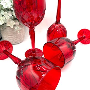 May include: Four red crystal wine glasses, two lying on their sides, with intricate cut patterns. One glass stands upright, and another is in the background. The stemware is ruby red, reflecting light, and is set against a white background.