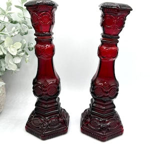 May include: A pair of deep red glass candle holders featuring a pressed pattern with circular and geometric motifs. Each candlestick has a square base, a tapered stem, and a wide top rim, showcasing a translucent finish and intricate detailing.