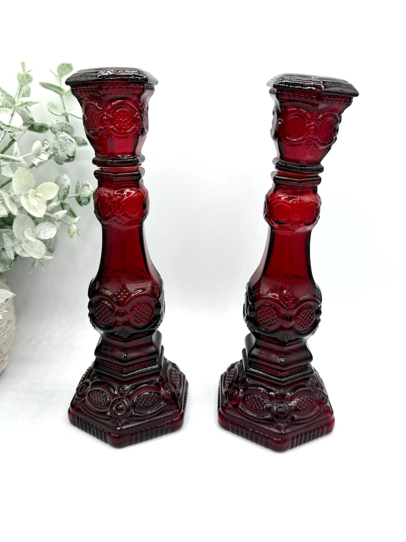 May include: A pair of deep red glass candle holders featuring a pressed pattern with circular and geometric motifs. Each candlestick has a square base, a tapered stem, and a wide top rim, showcasing a translucent finish and intricate detailing.