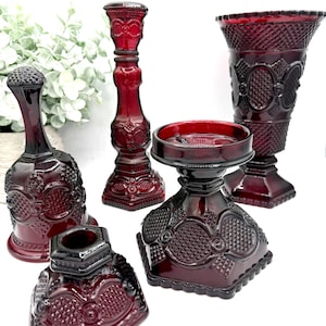 May include: A collection of ruby red glass decorative items, including a bell, a candlestick, a candle holder, and a vase. The pieces have ornate, textured designs and are arranged on a white surface. The tallest piece is approximately 8 inches tall.