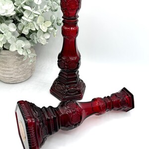 May include: Two deep red glass candle holders featuring an intricate pressed pattern with geometric shapes and circular motifs. One candle holder stands upright while the other lies horizontally, both showcasing a hexagonal base and neck.