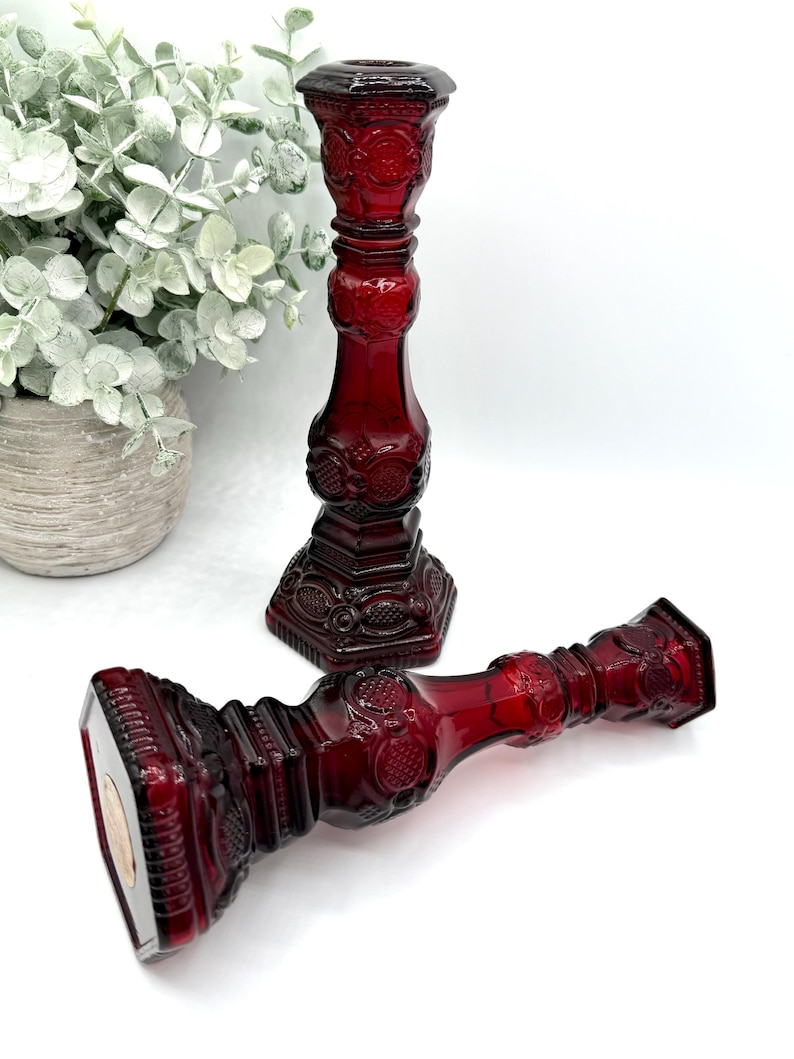 May include: Two deep red glass candle holders featuring an intricate pressed pattern with geometric shapes and circular motifs. One candle holder stands upright while the other lies horizontally, both showcasing a hexagonal base and neck.