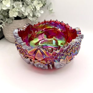 May include: A round, iridescent carnival glass bowl featuring a vibrant red and gold finish with a complex geometric diamond pattern and a serrated, sawtooth rim. The bowl displays a reflective, multi-colored sheen across its textured surface.
