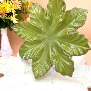 May include: A glossy, green ceramic leaf-shaped serving dish. The dish has a detailed, veined texture and a slightly darker rim. It is displayed on a clear stand, with a vase of flowers in the background.