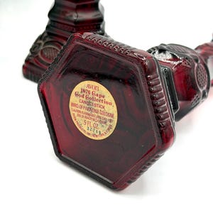 May include: Two deep red glass bottles shaped like candlesticks with textured geometric patterns. The base of one bottle features a circular label reading: AVON 1876 Cape Cod Collection CANDLESTICK BIRD OF PARADISE COLOGNE 5 FL. OZ. 32289.