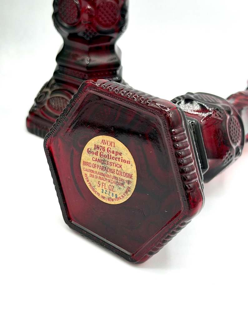 May include: Two deep red glass bottles shaped like candlesticks with textured geometric patterns. The base of one bottle features a circular label reading: AVON 1876 Cape Cod Collection CANDLESTICK BIRD OF PARADISE COLOGNE 5 FL. OZ. 32289.