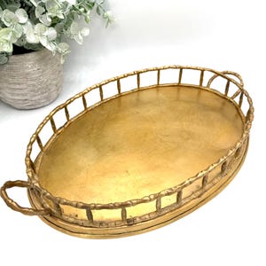 May include: An oval-shaped gold-toned metal tray featuring a raised gallery rail with a textured bamboo-style design. The tray includes two matching handles on the sides and a smooth, flat base, providing a functional surface for display.