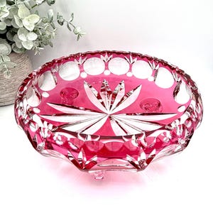 May include: A ruby red, oval-shaped crystal bowl with a starburst design in the center. The bowl features a cut glass pattern with circular and geometric shapes. The bowl is elevated on small feet and is likely used for decorative purposes.