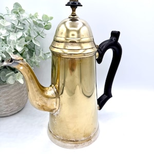 May include: A vintage brass coffee pot with a black handle and a decorative top. The pot has a curved spout and a cylindrical body. The brass has a warm, golden hue, and the handle is a glossy black.