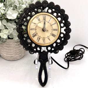 May include: A black cast iron electric clock with a round, ornate, scalloped frame and a handle at the bottom. The clock face is cream-colored with black Roman numerals, gold-toned trim, and two black hands. A black power cord is attached to the back.