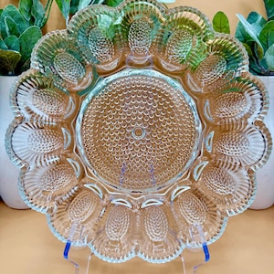 May include: A clear glass egg serving platter with a scalloped edge and individual egg-shaped compartments. The center of the platter has a textured, dotted design. The platter is displayed on a clear plastic stand, with green plants in the background.